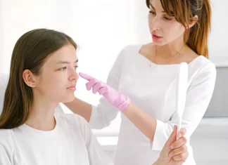 Dermatolog w rękawiczkach bada policzek pacjentki trzymającej lusterko.