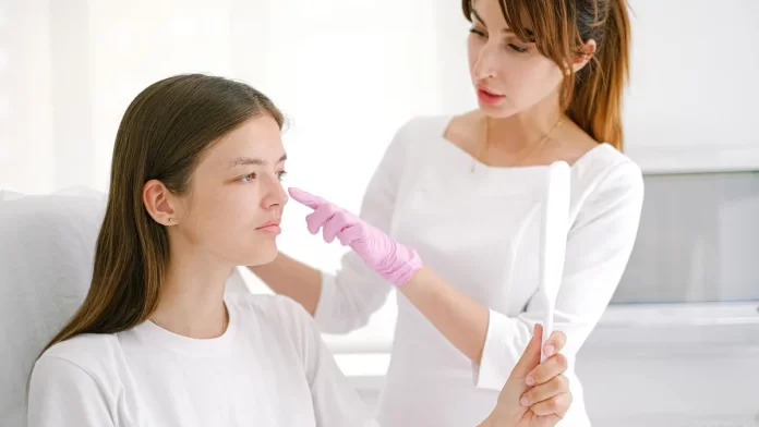 konsultacja-dermatologiczna-problem-naczynek Dermatolog w rękawiczkach bada policzek pacjentki trzymającej lusterko.